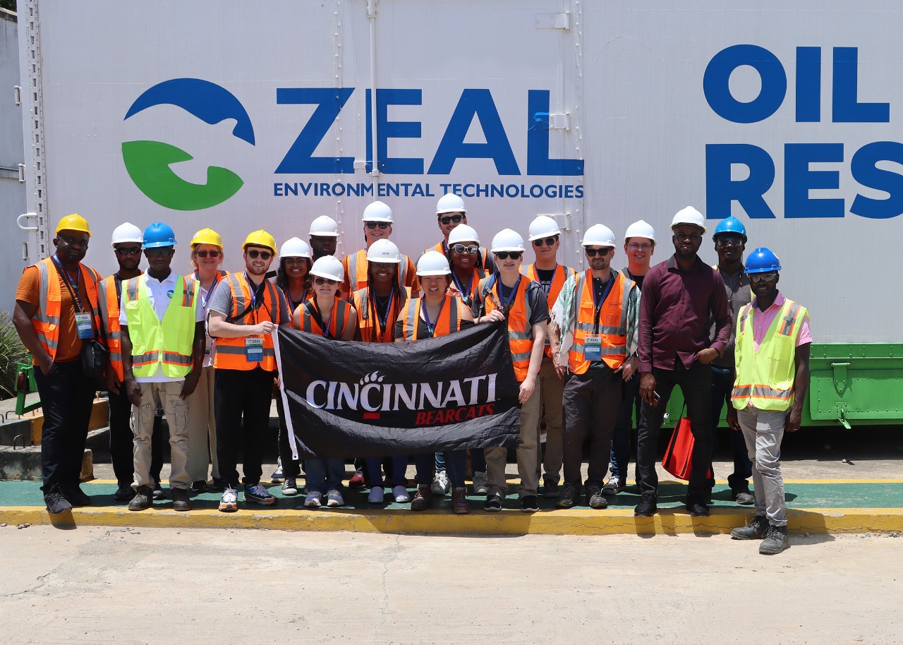 A group wearing hard hats and high-vis safety vests poses with a banner that reads “Cincinnati Bearcats” in front of a large truck that has “Zeal Environmental Technologies” painted on it; also visible on the truck are the word “oil” and part of a word beginning with “res…“