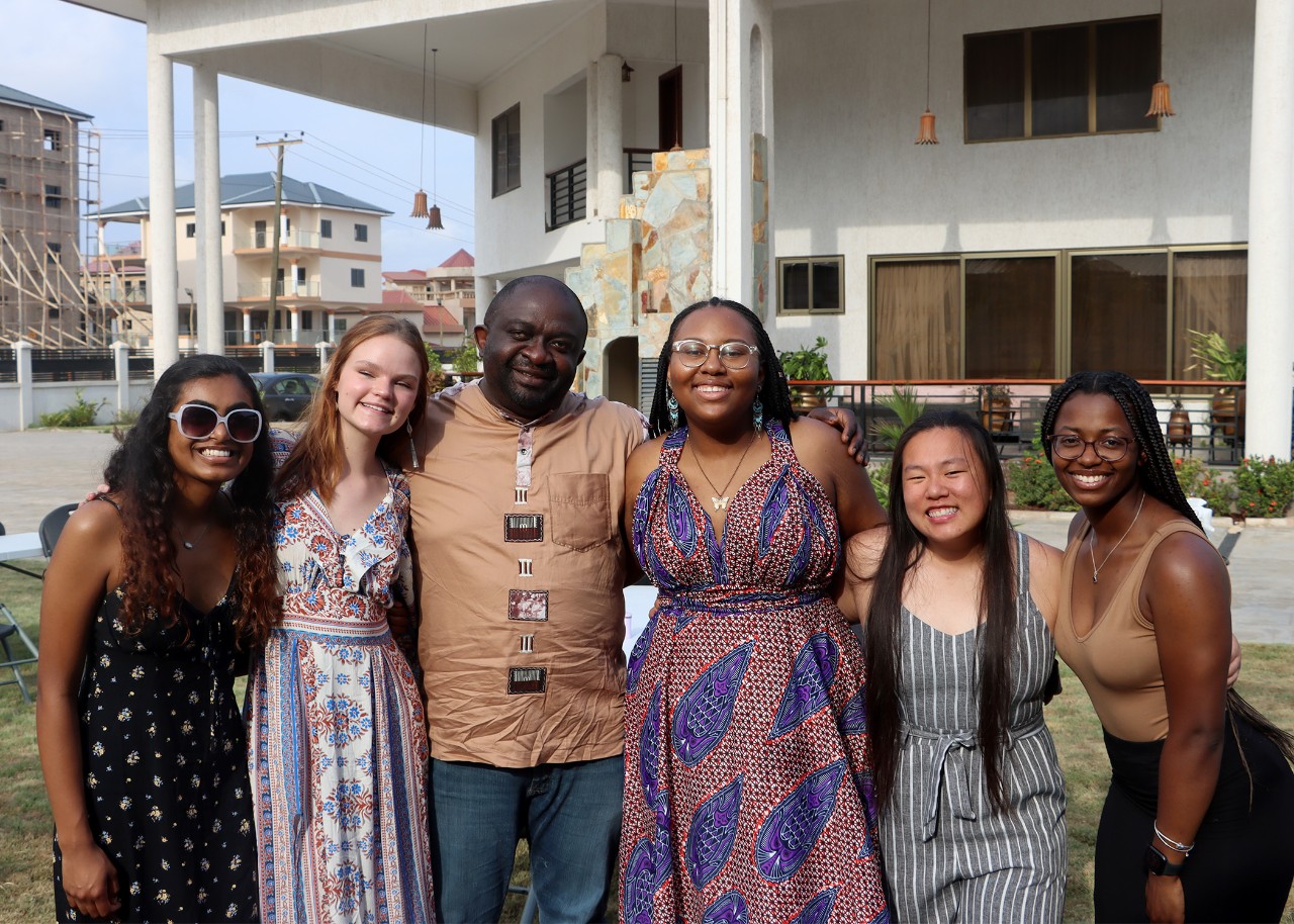 A row of people pose for the photographer on the grass in front of a white building; other buildings, one with wooden scaffolding, are behind them