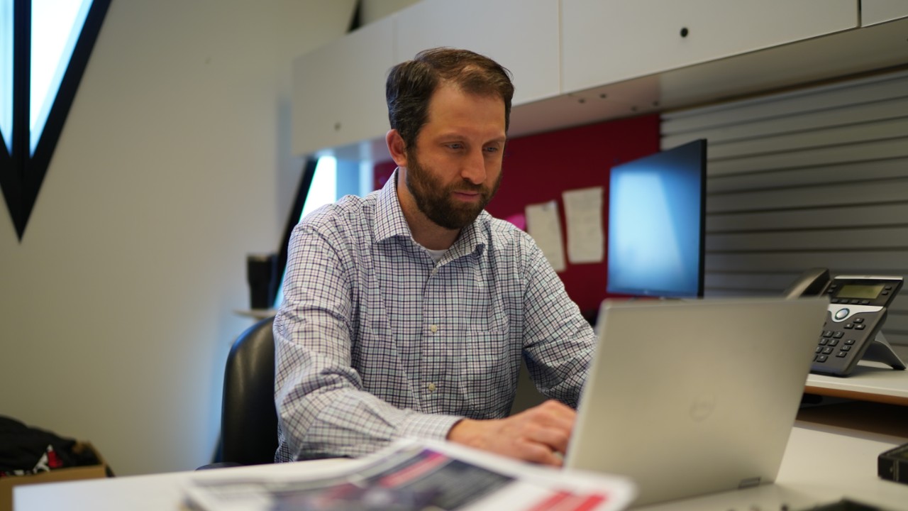a photo of a bearded man seated at his desk working on his laptop