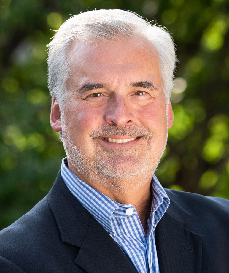 A man in a blue shirt and a dark blue jacket smiles.