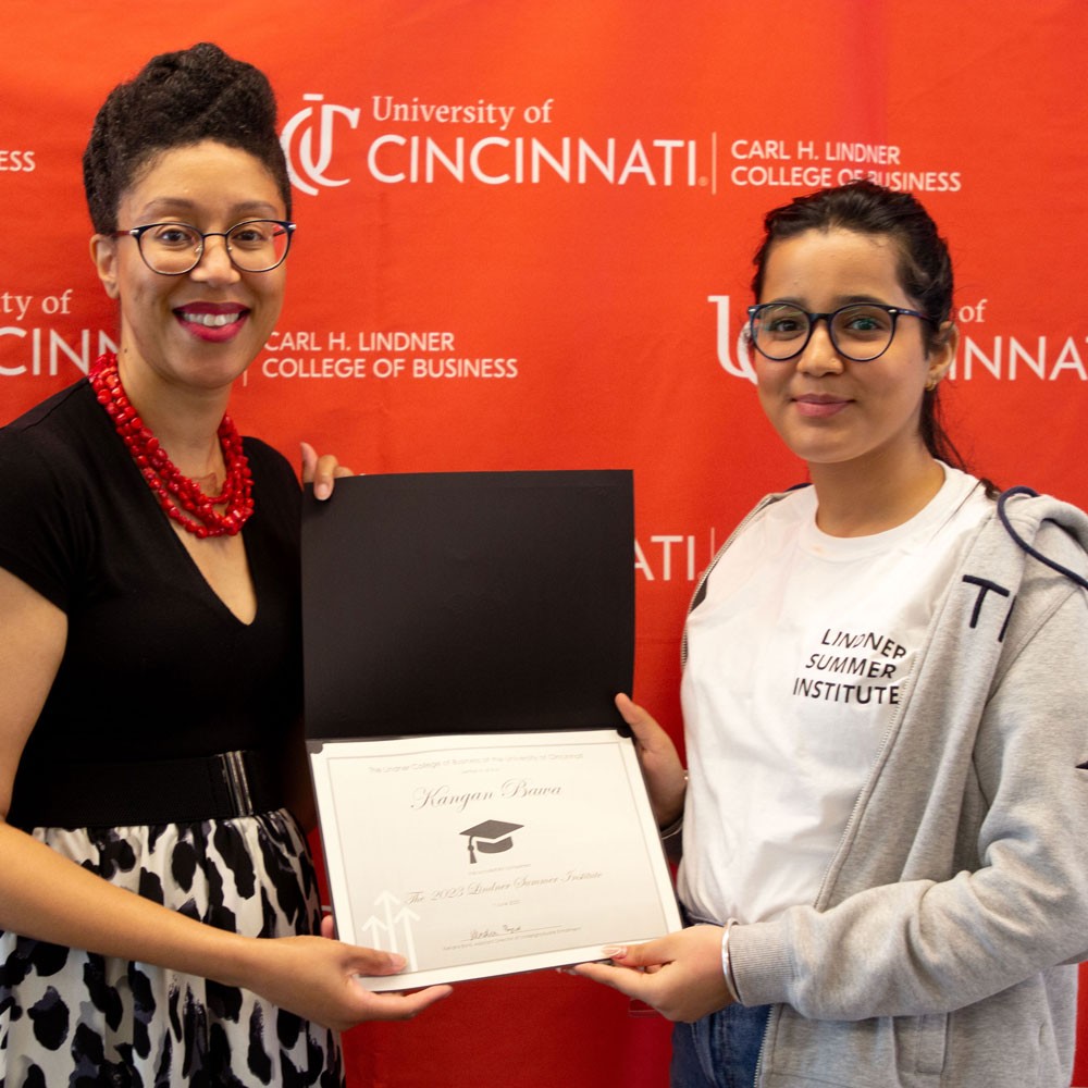 woman wearing black top and red necklace and glasses presents white certificate in black folder to a female high school student wearing a white shirt and grey sweater and glasses against a red backdrop
