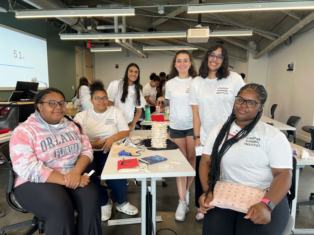 six female high school students sit or stand around their end of a long white rectangular table and their completed tabletop lamp made our of clothespins and glue