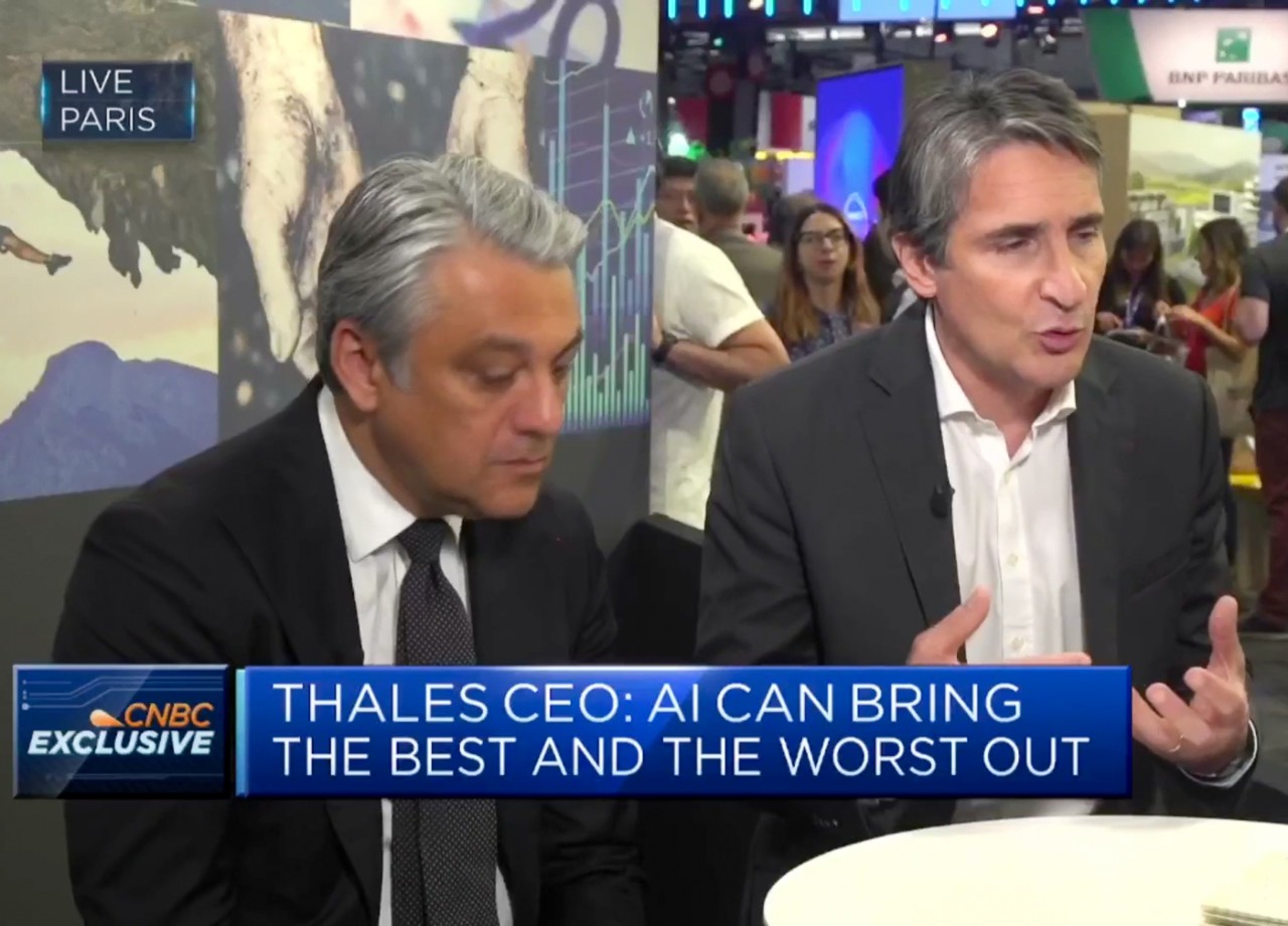 Two men are interviewed on a convention floor with people walking in the background. 