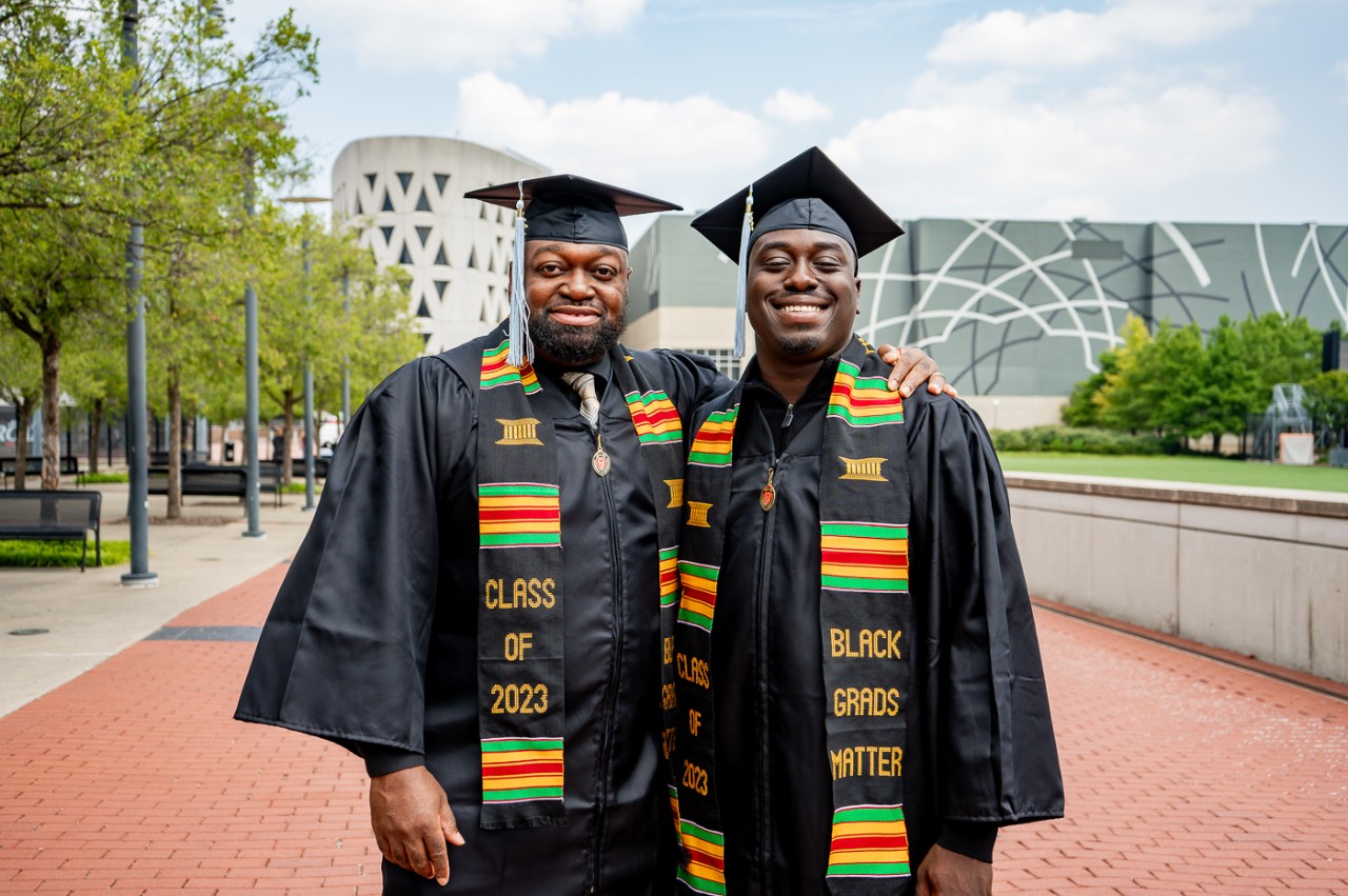 Two grads in caps and gowns pose on UC's Uptown Campus.