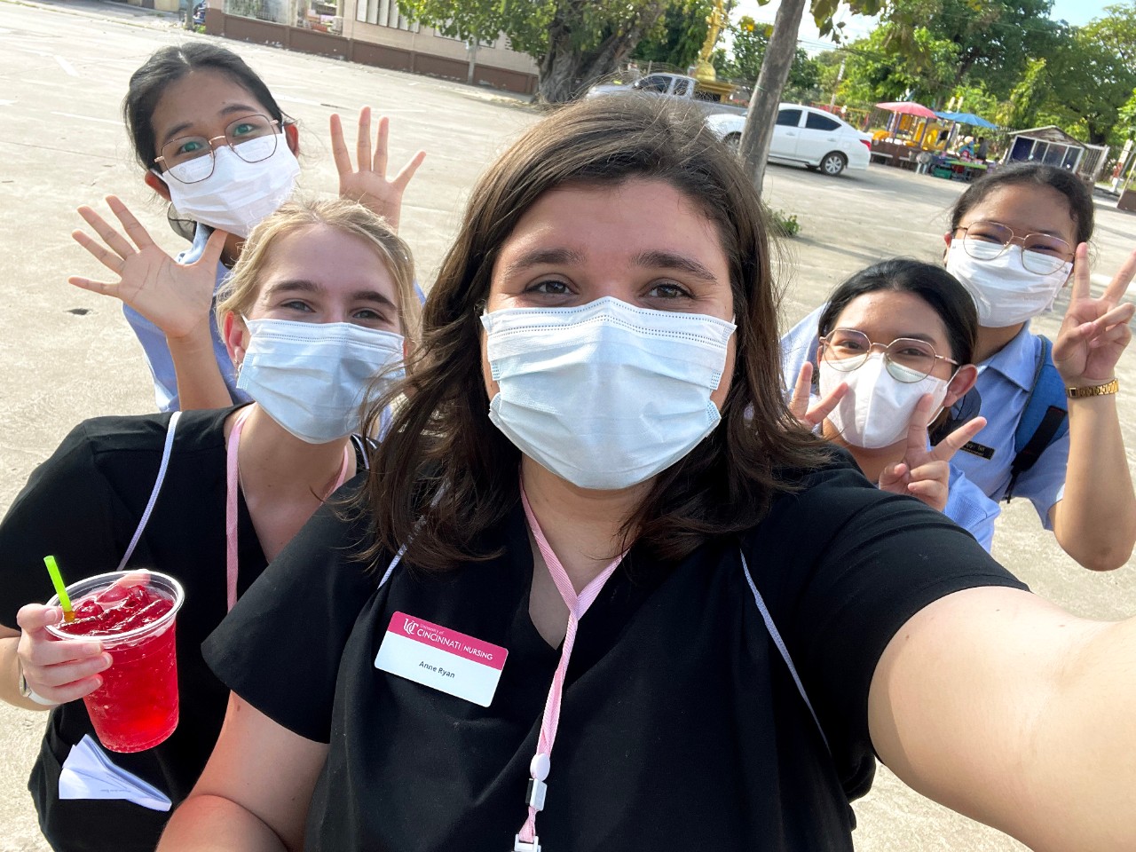 A small group of young women pose masked for a selfie in a parking lot, smiling and gesturing for the camera; the woman in front wears a nametag reading University of Cincinnati Nursing, Anne Ryan.