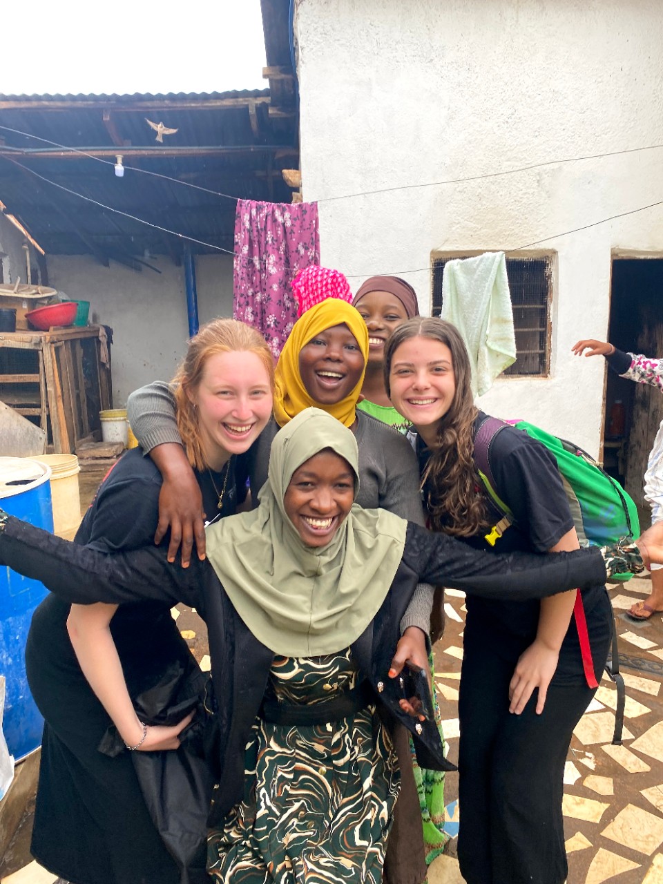 Five smiling young women, some Black and wearing hijab, some white and wearing black t-shirts and skirts, lean closely together for a photo in what looks like a small courtyard; the young woman in front spreads her arms widely, and behind is a white stucco building with a tin-roofed lean-to, some barrels, and some things hanging on a clothesline.