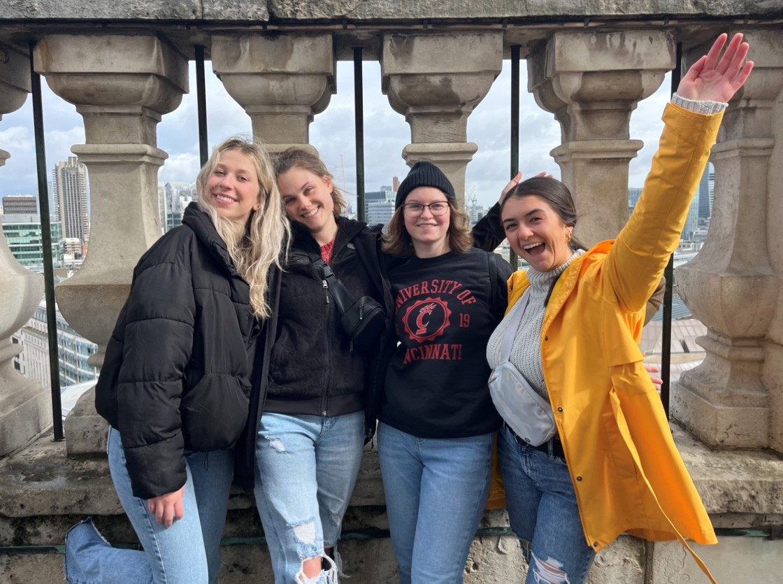 Four young women post in front of a large stone balustrade; one wears a bright yellow rain jacket and another a University of Cincinnati sweatshirt.