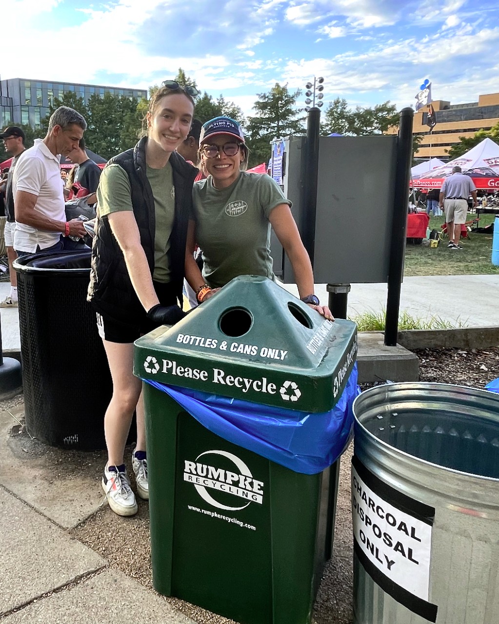 Two women stand next to a green recycle can on UC's Sigma Sigma Commons on football game day.