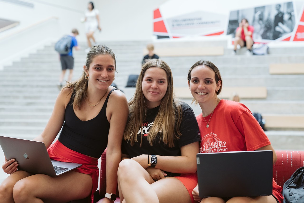 Three students in red and black clothing sit in the Lindner Hall atrium.