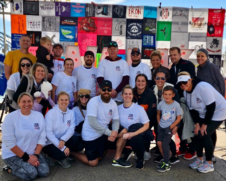 Group of people stand in front of a large quilt made of 'Walk Ahead' t-shirts.