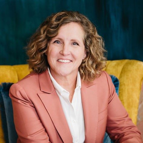 woman wearing pink blazer and white dress shirt sitting and smiling in a yellow chair with a blue background