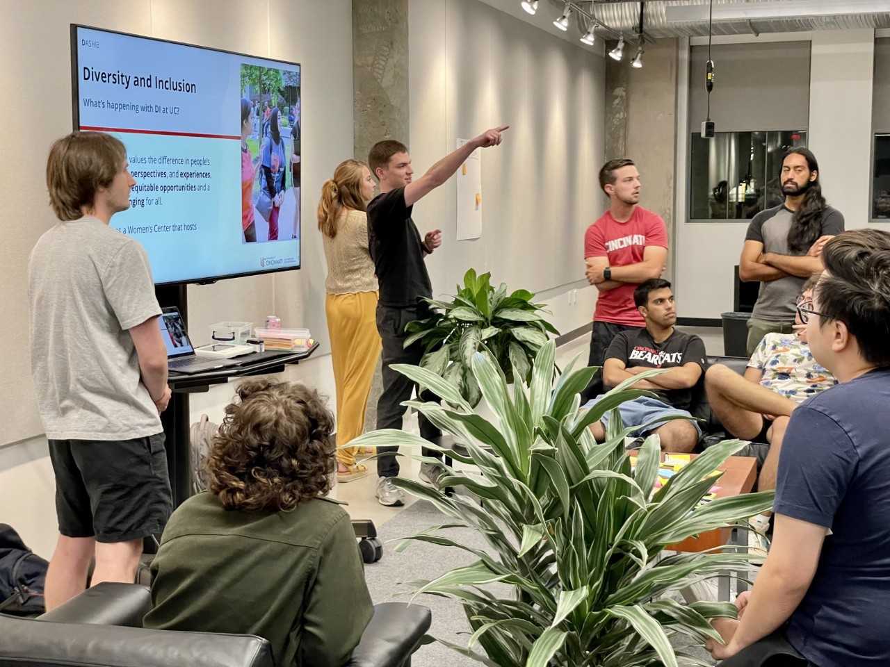 one students stands and is pointing while four students are also standing while two other students are sitting. There is a big screen which many in the room are watching.
