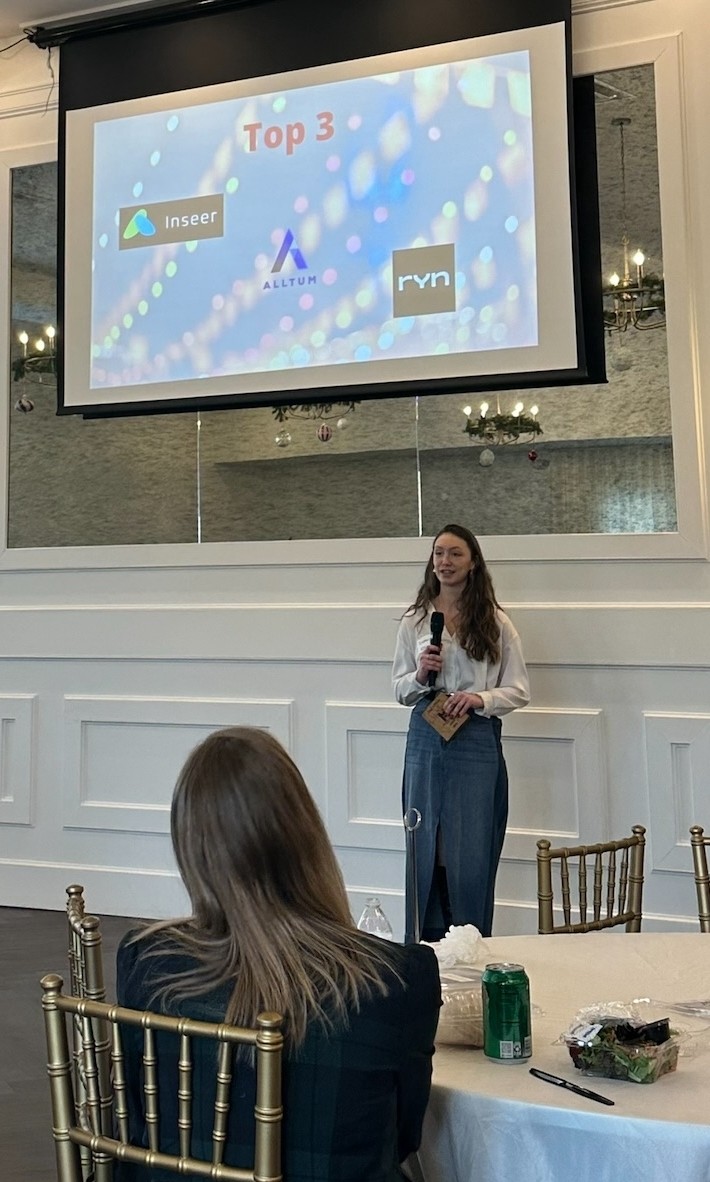 A woman at the front of a room gives a presentation to a group of seated individuals.