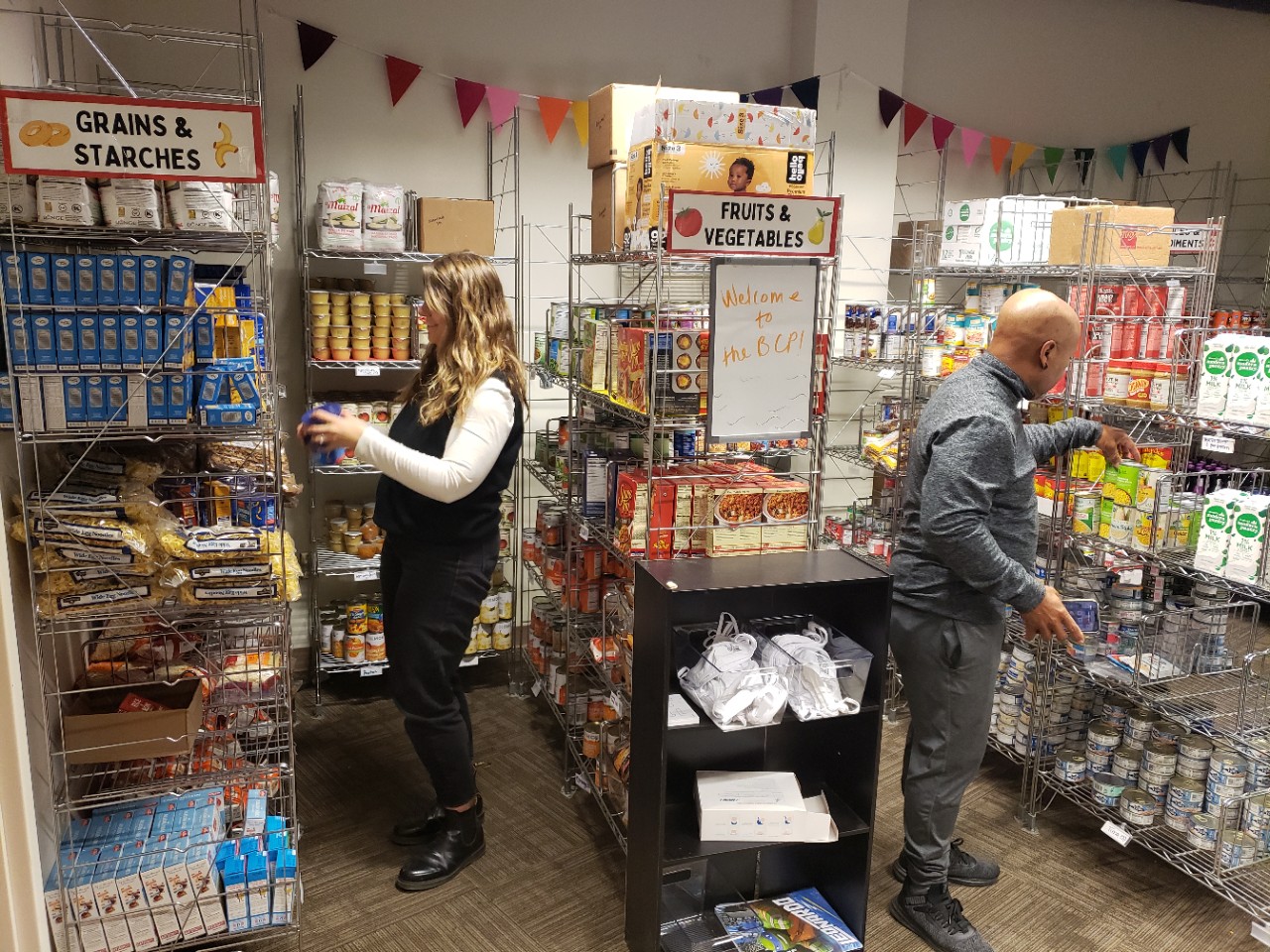 Three rows/columns of food items.  Joy Kostansek is shown stocking the first row and the second row shows Daniel Cummins stocking food items.
