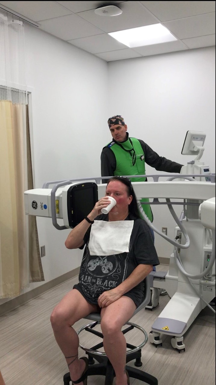 A patient sits and drinks from a cup while a technician takes an x-ray