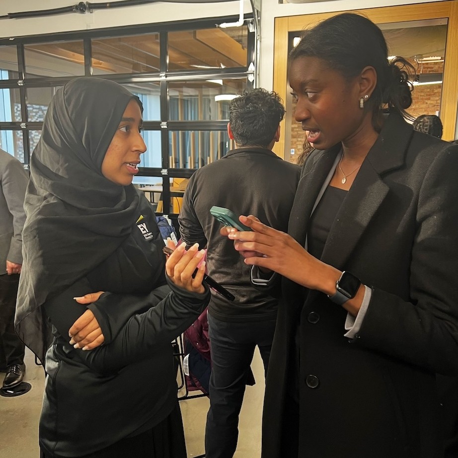 one woman wearing all black and a headscarf holds up a phone and talks to another woman in a black business suit in a large corporate meeting room