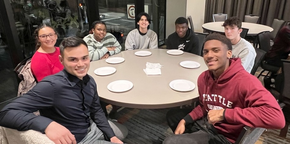 seven people sit at a round table in a hotel meeting room wearing casual clothing