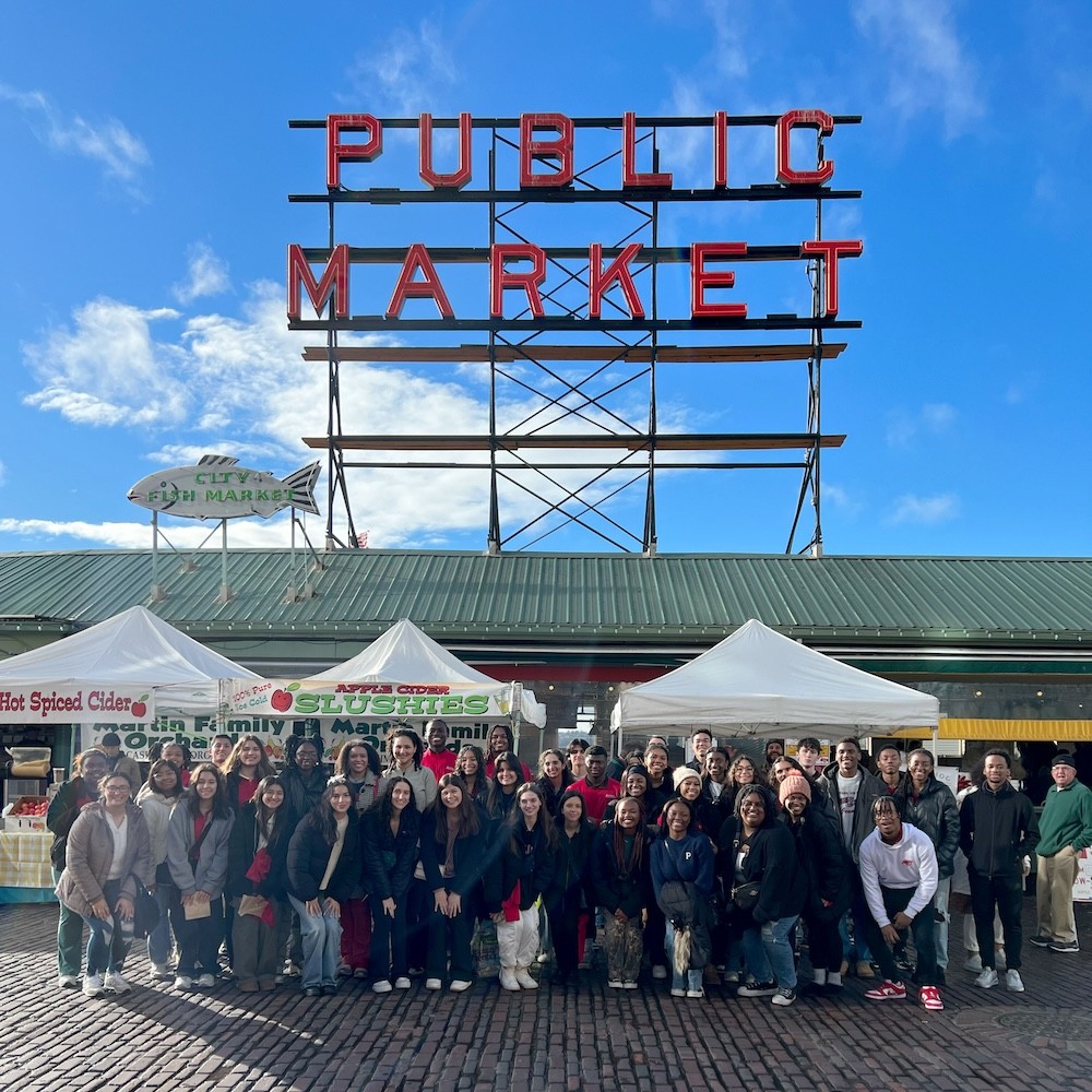 Nearly 50 students wearing sweats and outerwear stand and smile in front of three stalls with white tents and a large green roof with PUBLIC MARKET sign above them on a sunny day