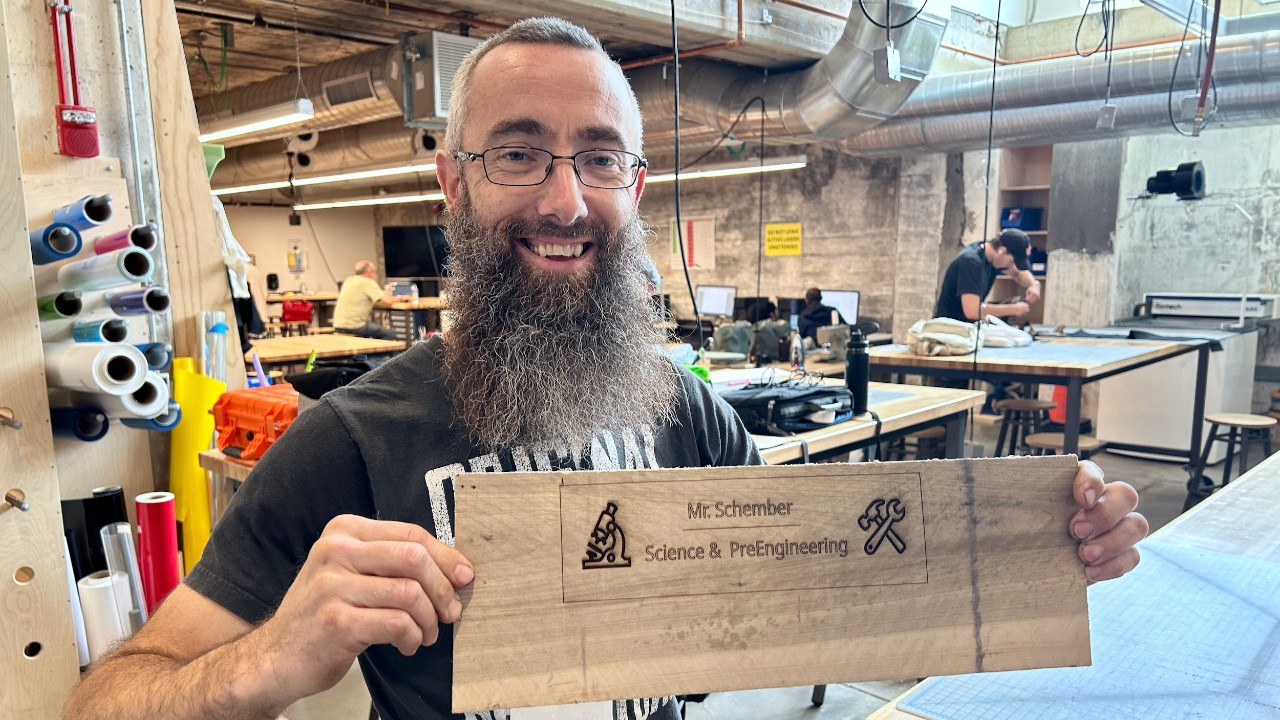A man hold up a wooden sign with his name engraved from a wood-working machine in UC's Makerspace.