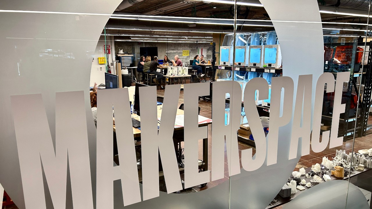 UC's Makerspace window sign with a view of people sitting at a table in the distance.