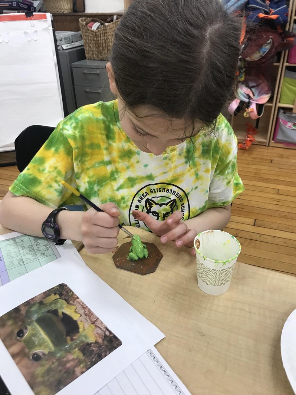 A child wearing a Clifton Area Neighborhood School T-shirt paints a frog green.
