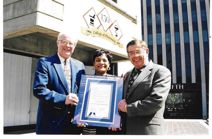 Two men and a woman hold a plaque in front of the College of Pharmacy building
