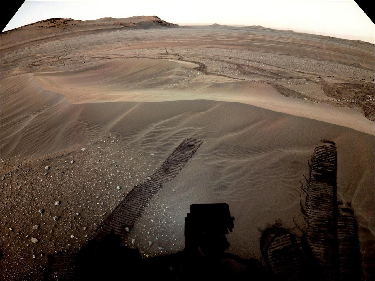 A wide shot of the sandy, hilly landscape of Mars with the tracks of the rover in the sand in the foreground and a bleached-out sky.