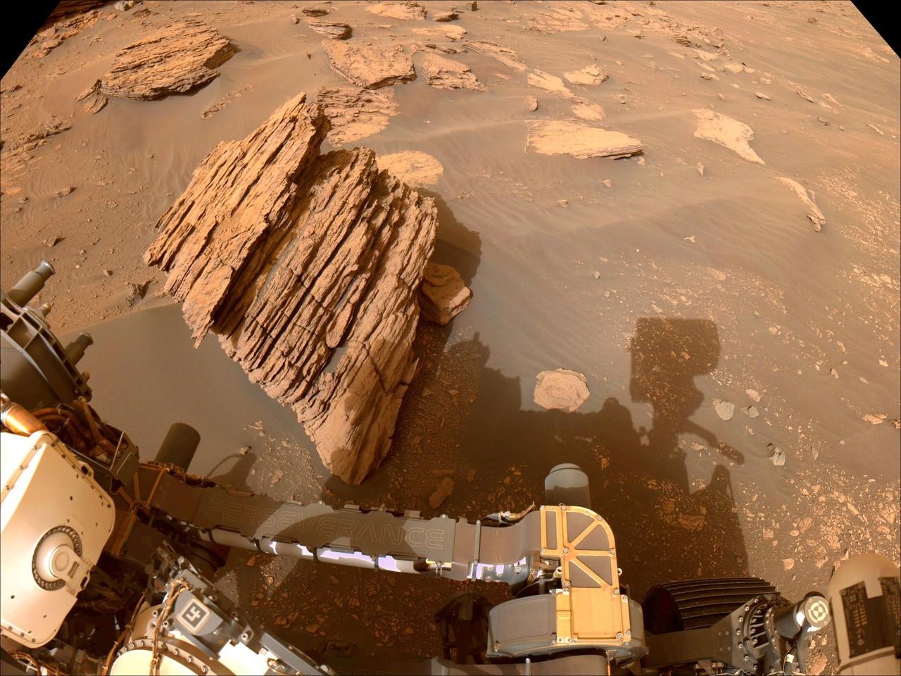The rover captures its own shadow across a striated boulder sticking up from the sand.