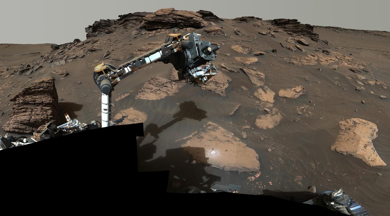 A close look at a rover's extended robotic arm with a background of Martian rocks and sand.