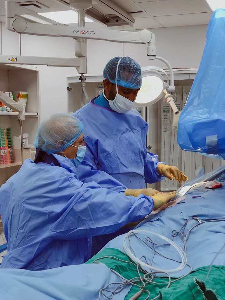 Attari and Meshesha, gowned and gloved, stand over a person draped for surgery; Attari’s hand is poised over the surgical field where Meshesha is working 