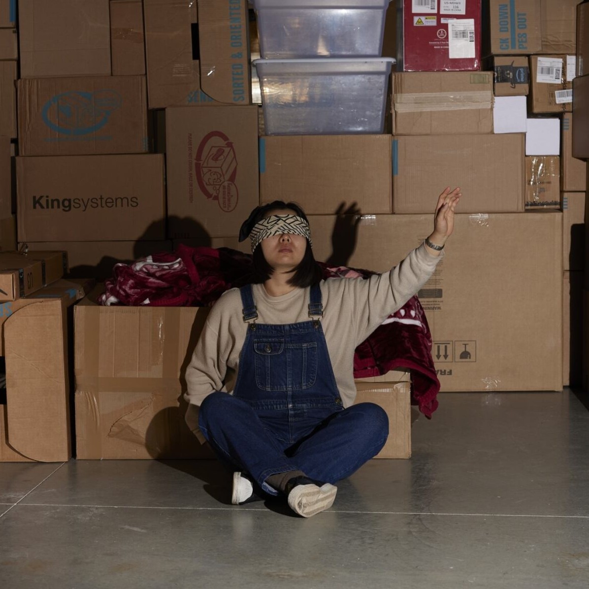 Photograph of a female wearing jean overalls sitting crisscross. She is blindfolded. She has a hand raised lightly. She is surrounded by boxes. She appears to be in a garage or storage unit.