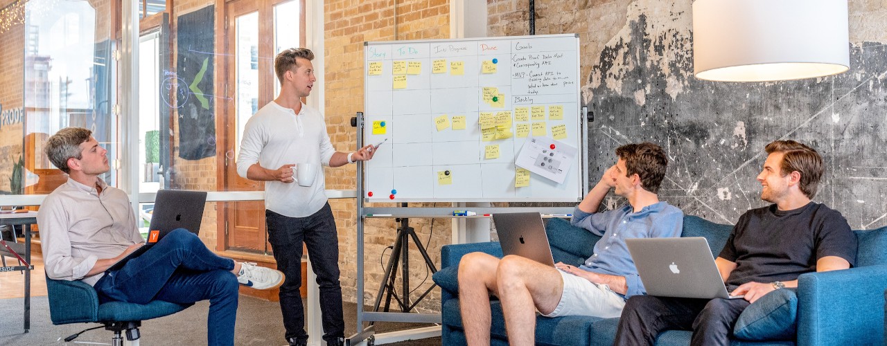 Three men sit in an innovation hub meeting while one man stands at a white board.