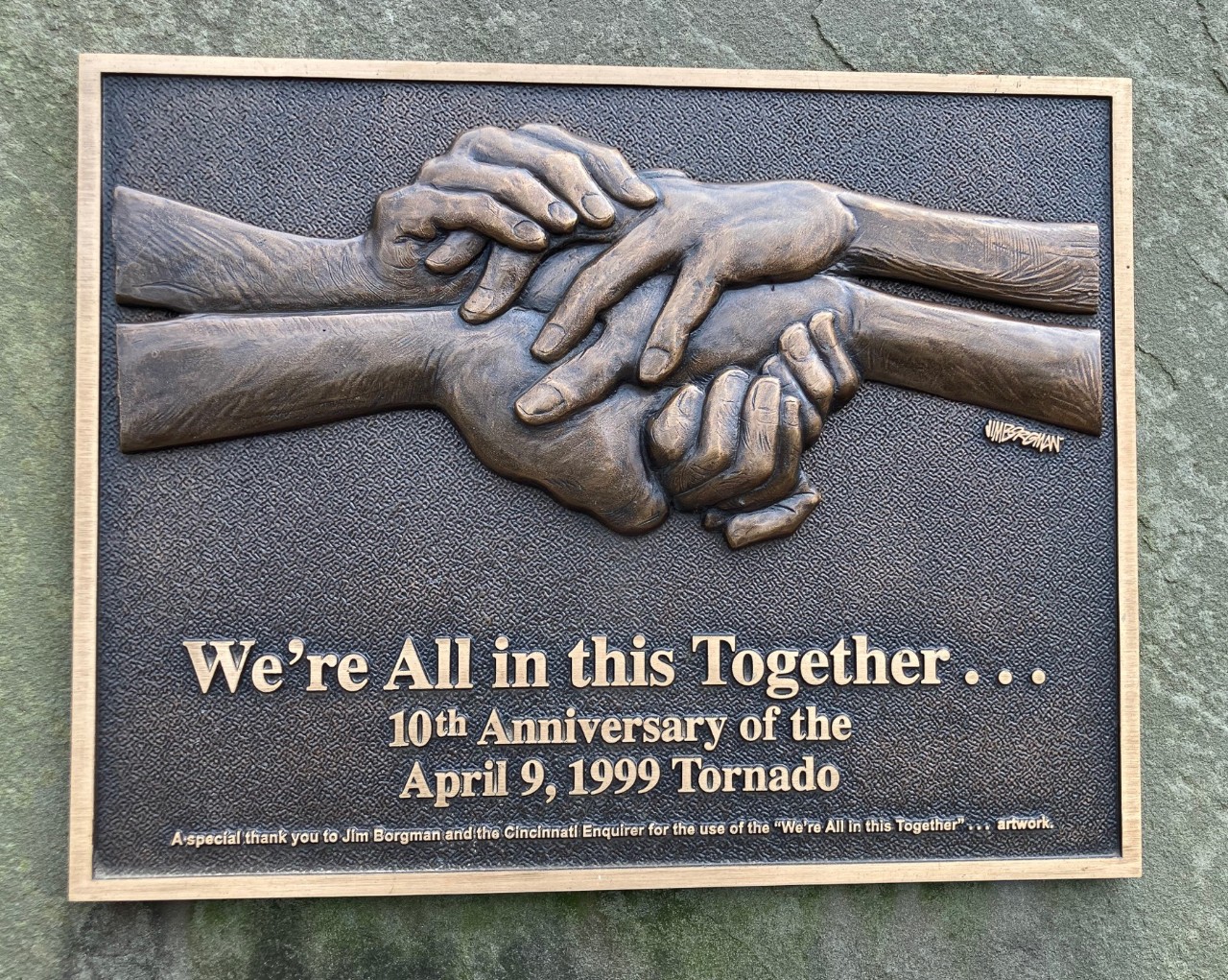 A bronze plaque features an bas relief of clasping hands above the words 'We're all in this together. 10th anniversary of the April 9, 1999 tornado."
