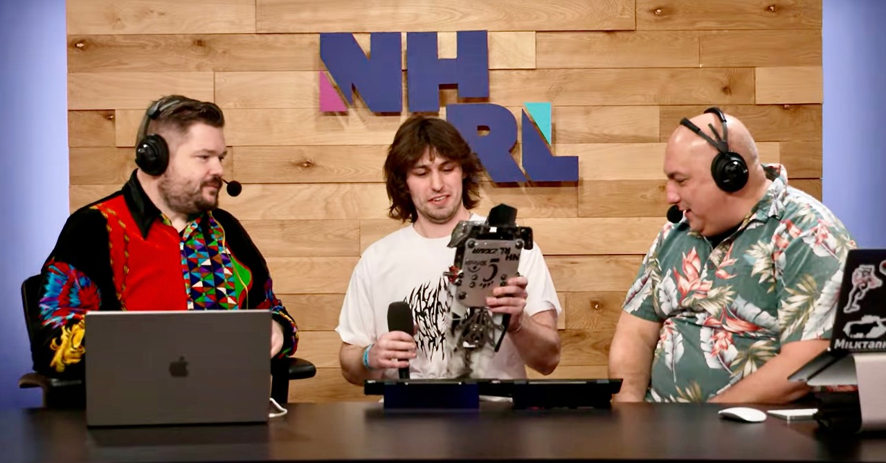 Three men sit together at a broadcasting desk discussing a combat robot held by Jacob Hoffmann in center.