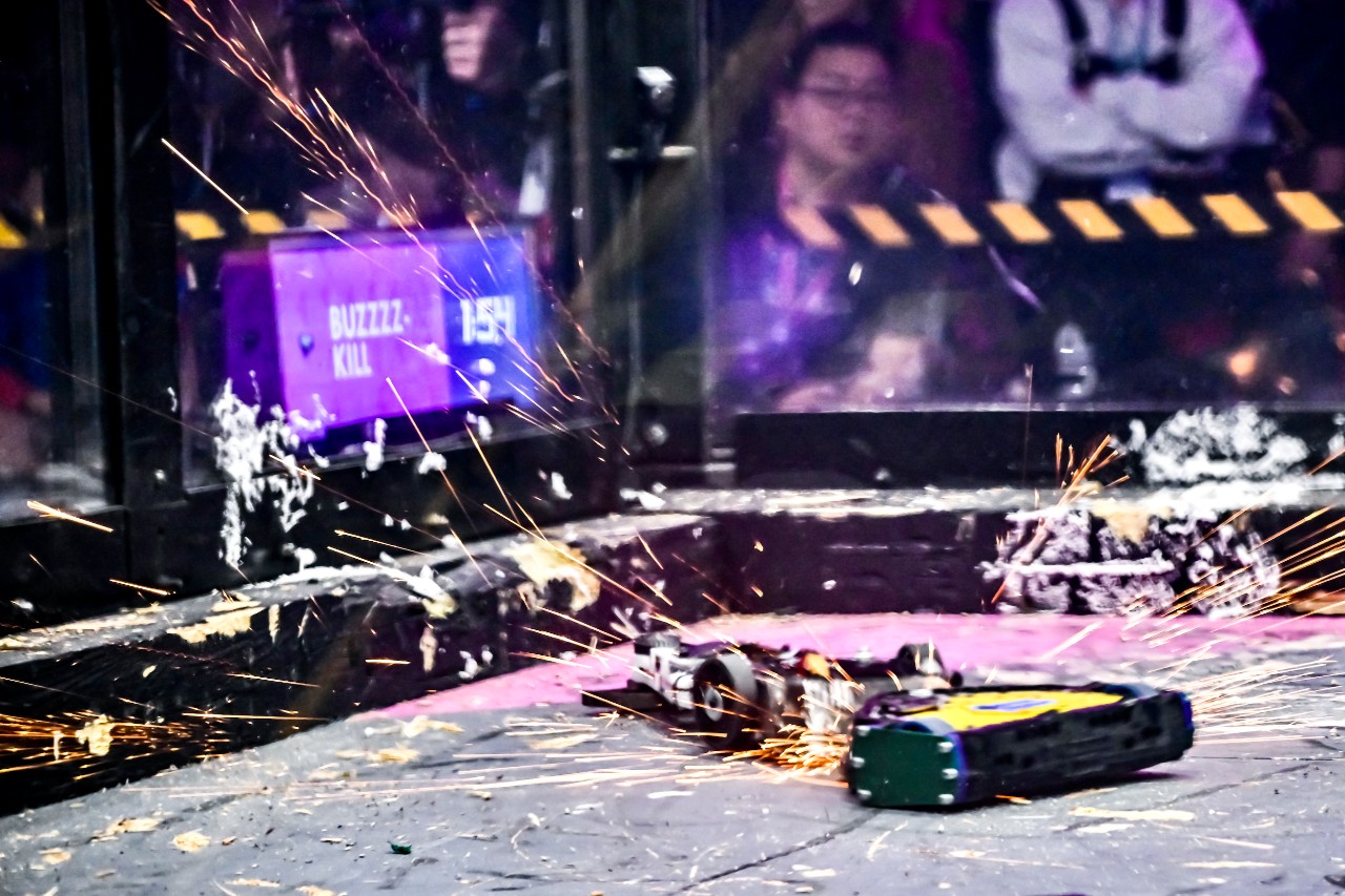 Two 12-pound combat robots face off inside a glass-walled enclosure shooting sparks and flames during contact.