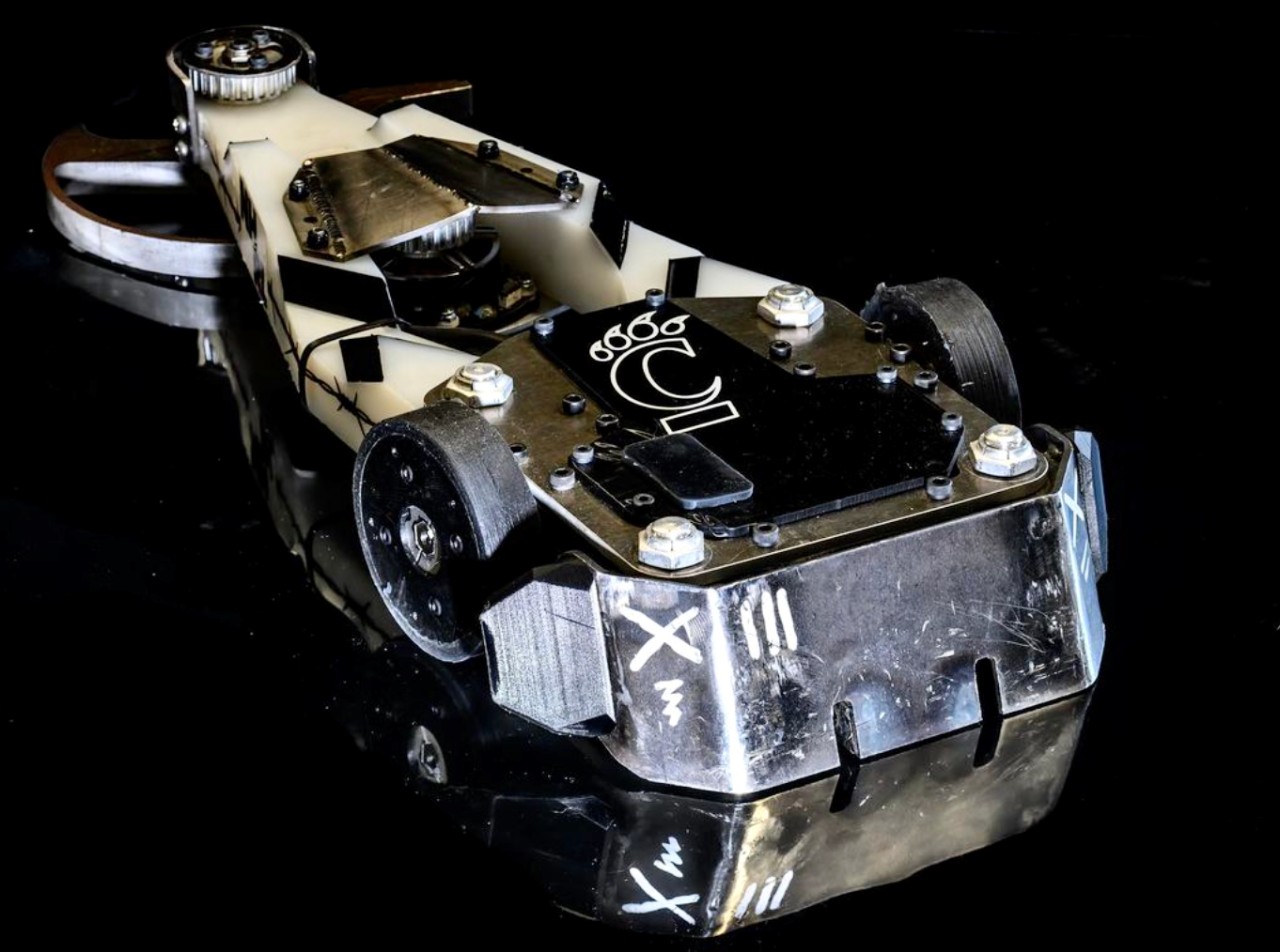 Close-up of a sliver combat robot that looks like a 12-pound Hotwheel car with a UC C-paw emblem on top.