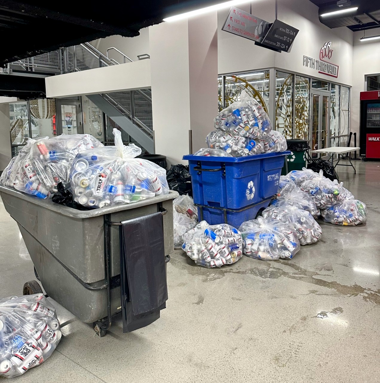 Twp large rolling garbage bins and several clear bags of recyclable cans in lobby of UC's Fifth Third Arena.