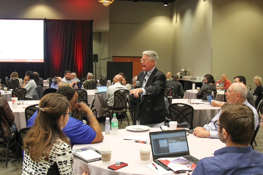 a man holding a microphone stands in front of a banquet table addressing a room full of attendees