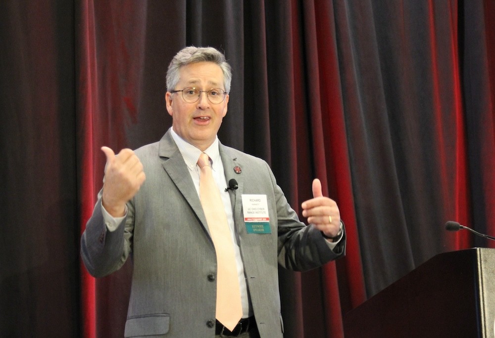 man in a grey suit and yellow tie speaks on a stage next to a podium in front of a black curtain uplit with red