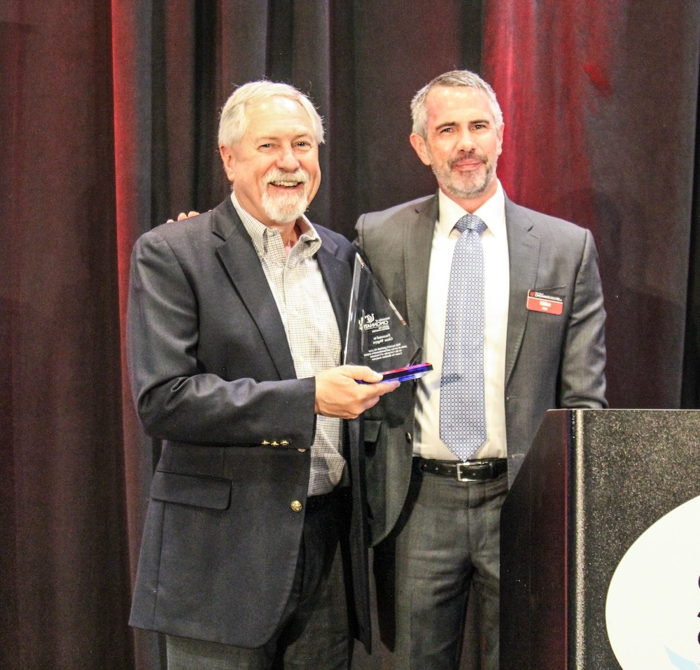 two men stand in business professional attire smiling and one of them holds a glass award