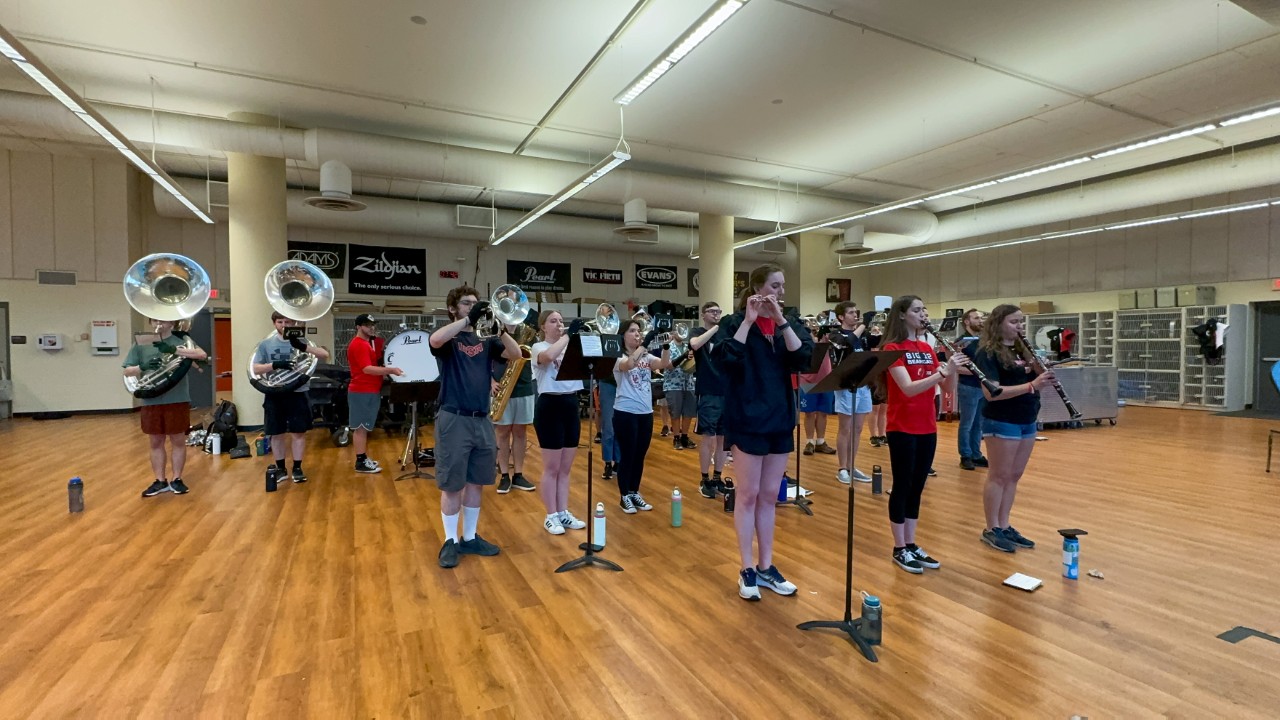 Members of a marching band stand and play music in a rehearsal room.