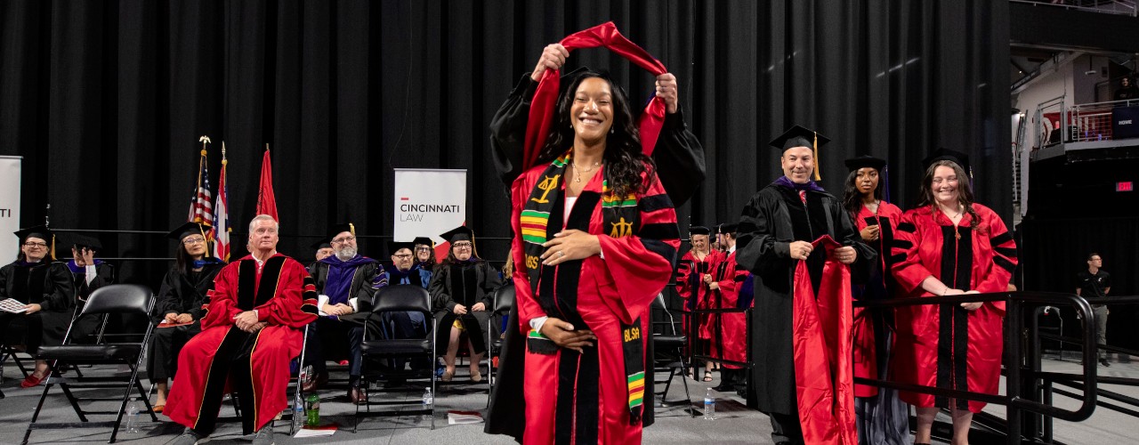Several faculty members dressed in doctoral regalia sit on a commencement stage as a woman front and center stands facing front as she is hooded at a law school graduation.