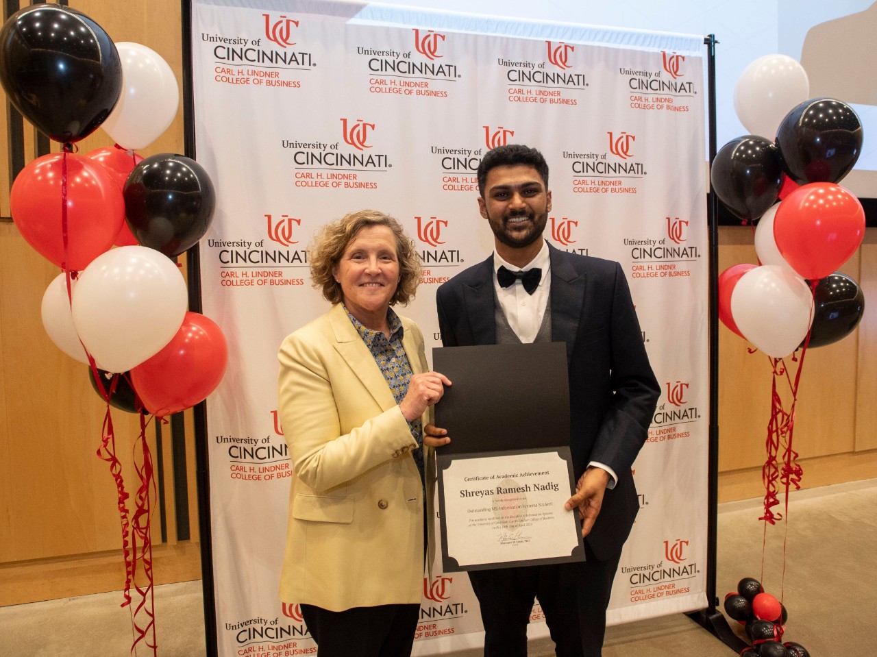 A woman in a tan suit jacket holds up a plaque with a student in professional dress.