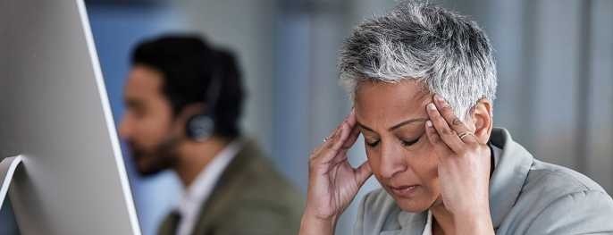 A woman sits at a desk in front of a computer with her hands on her temples and eyes closed and a pained expression on her face