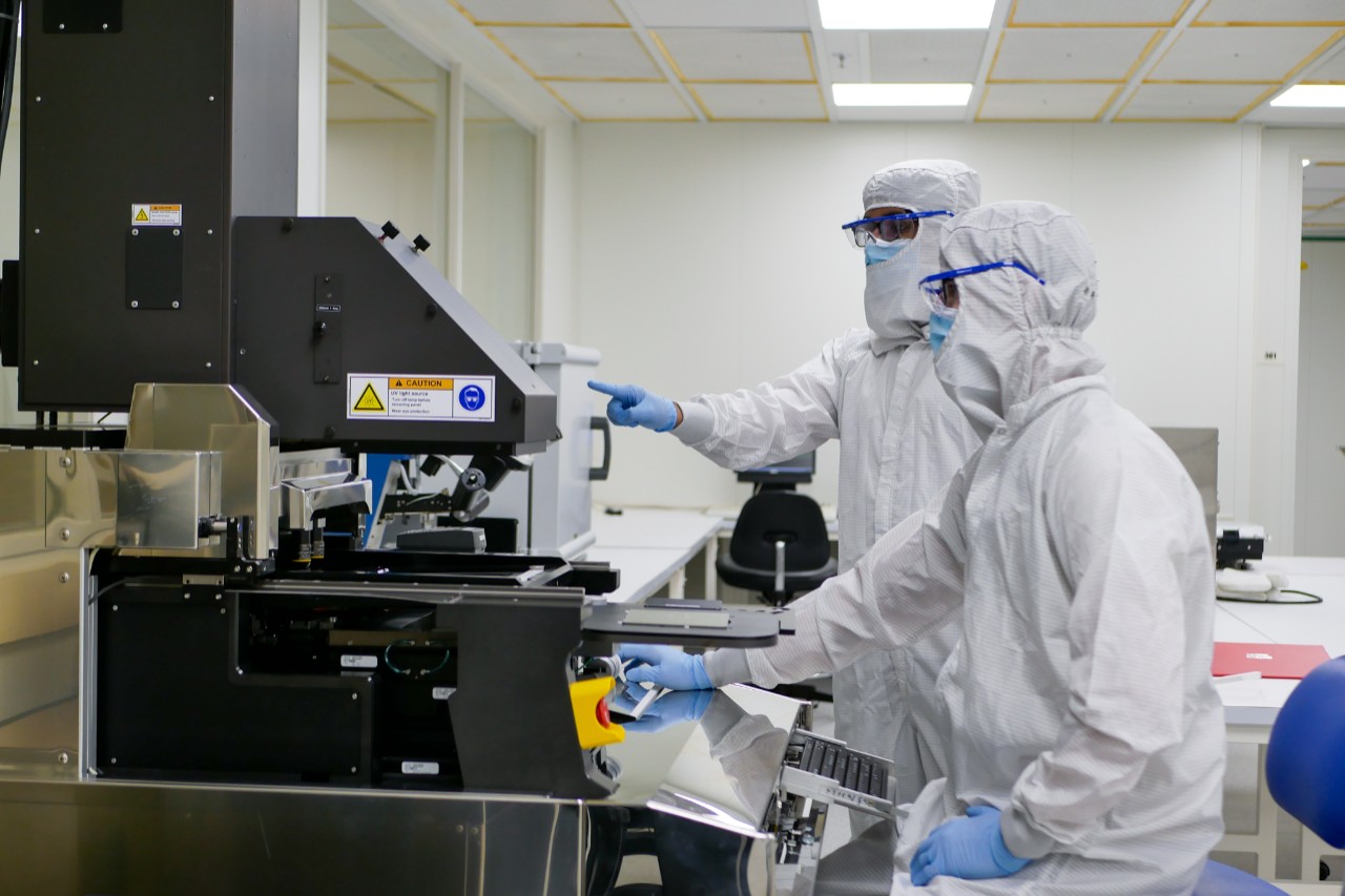 Two students in head-to-toe protective gear work at a machine in a clean room.