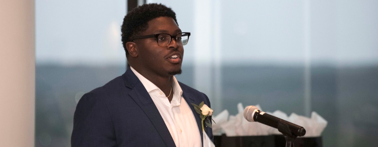 UC NIS students Lance Entsuah stands at a microphone wearing a white rose on his black suit jacket lapel.