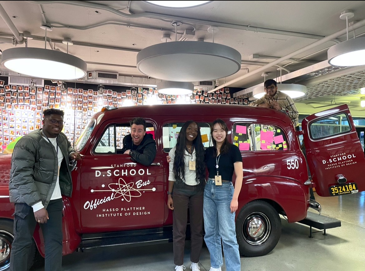 Four college students stand outside a maroon vintage automobile with a man sitting in the driver's seat.