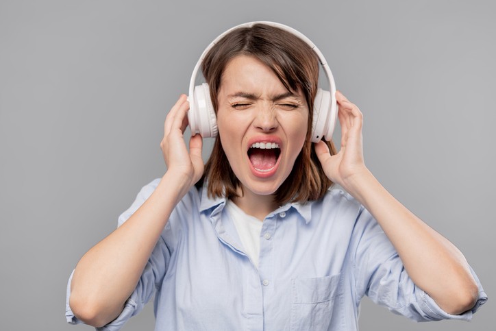 A woman wearing headphones holds her hands to her ears and yells with her eyes closed