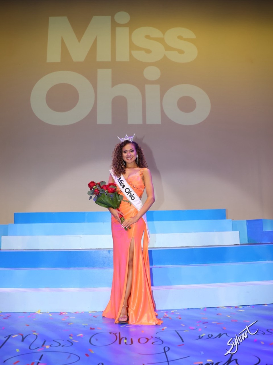 Miss Ohio onstage in orange dress, sash, crown, holding flowers