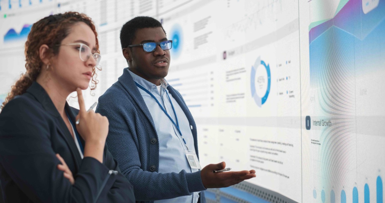 Two people dressed in business attire look at a large white screen with data on it.
